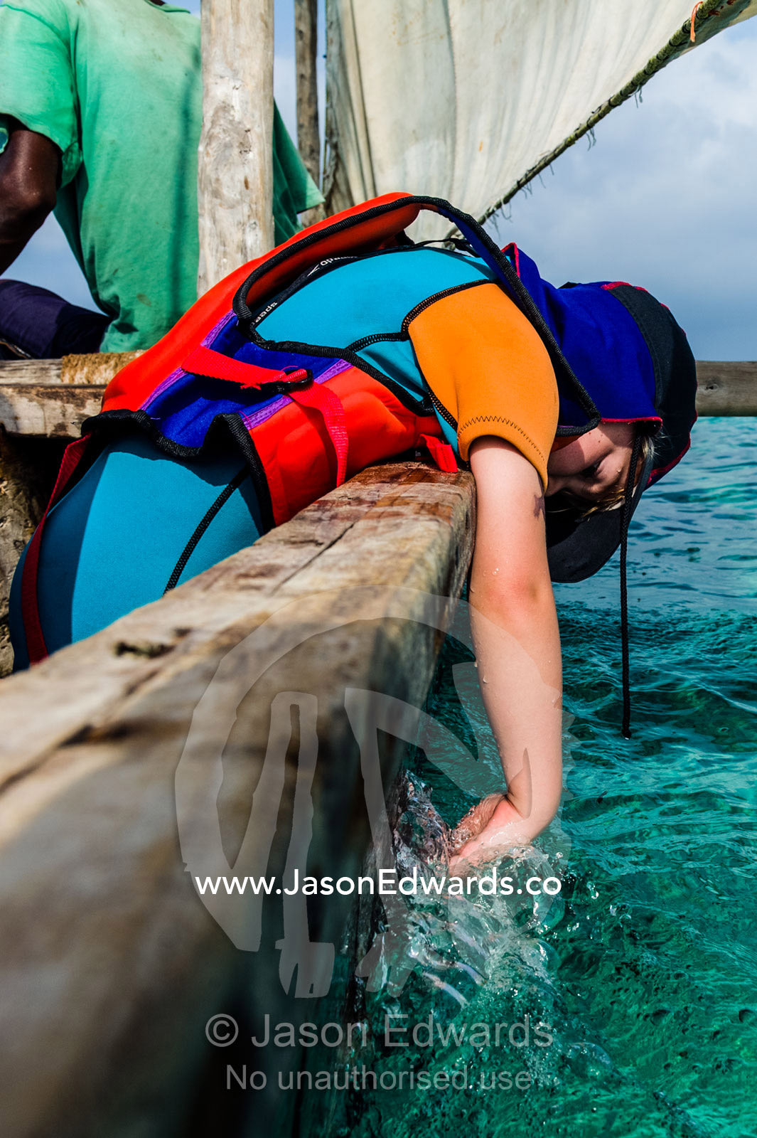 A small boy wearing a life jacket and wetsuit trails his fingers in the ocean from a wooden dhow. Bwejuu, Unguja Island, Zanzibar, Tanzania, Africa.
