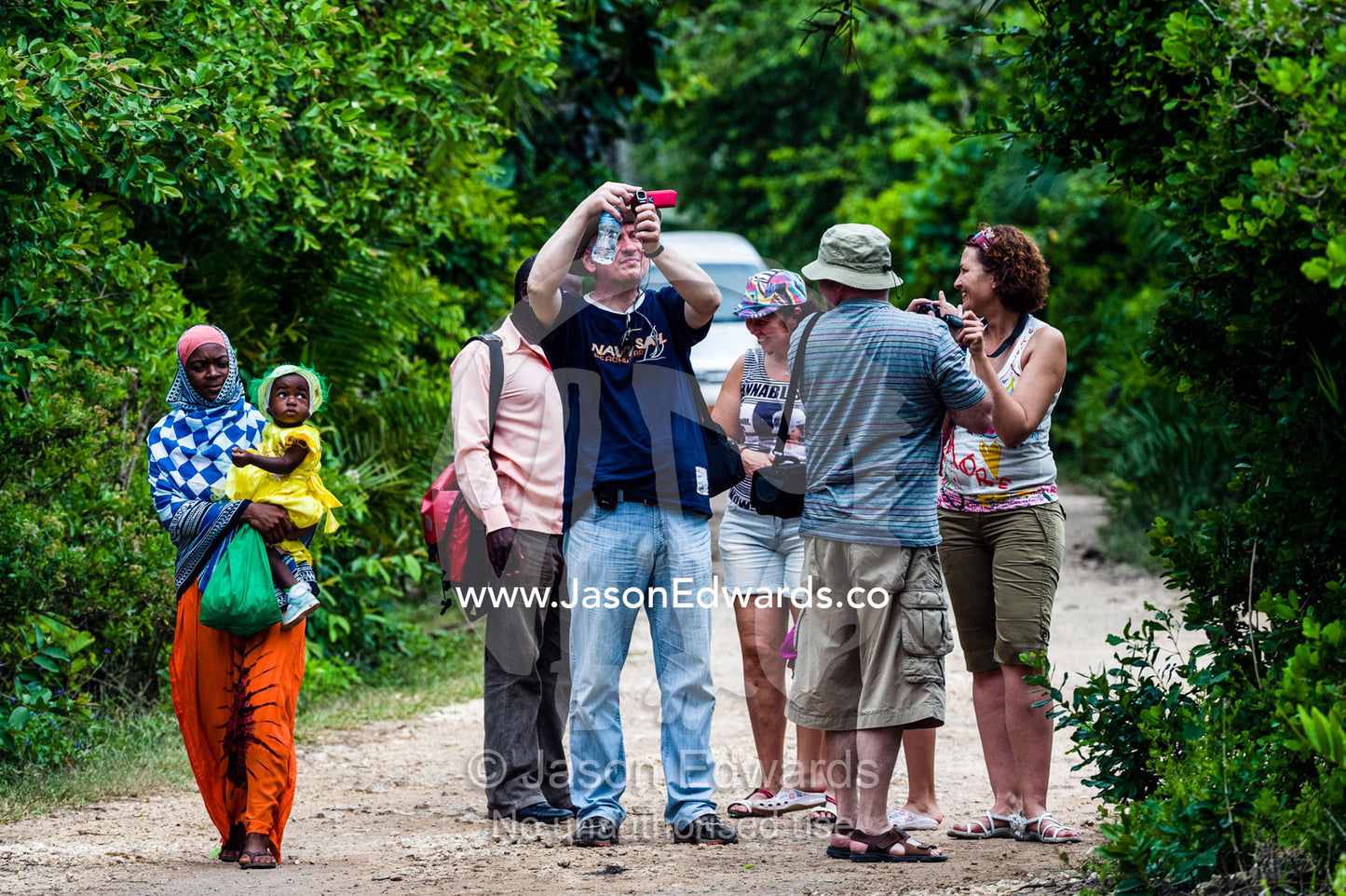 A villager and baby walk by tourists watching Zanzibar Red Colobus in a Coral Rag Forest. Ungula Island, Jozani Chwaka Bay National Park, Zanzibar, Tanzania, Africa.