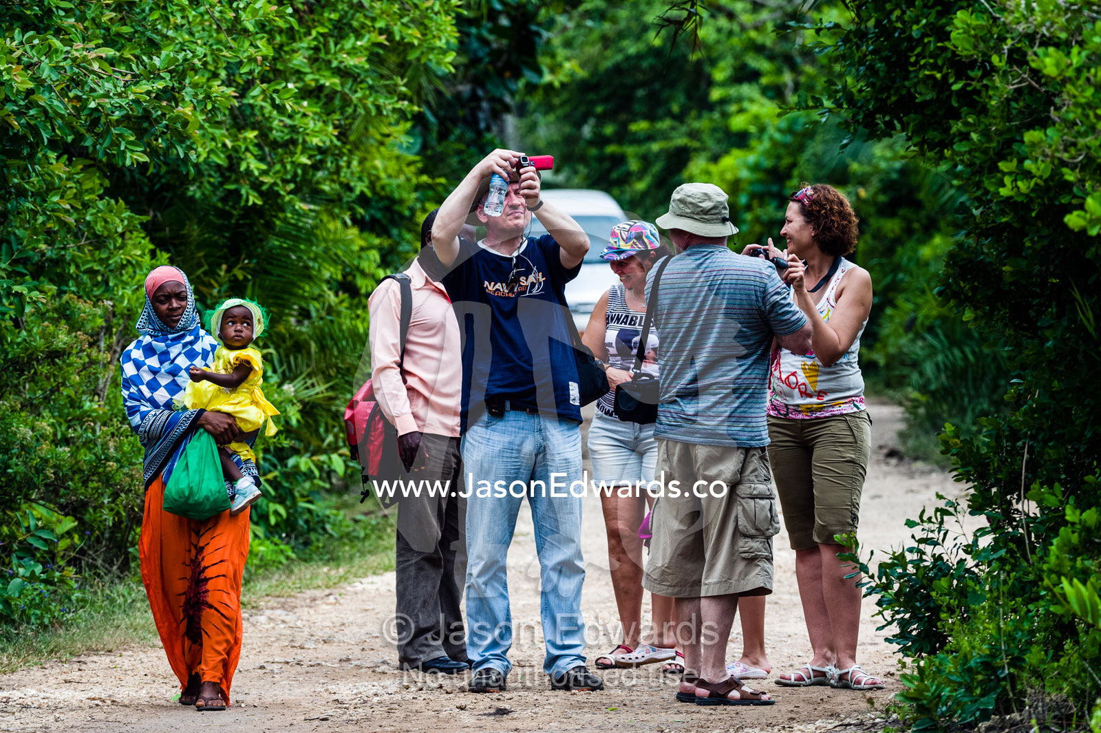 A villager and baby walk by tourists watching Zanzibar Red Colobus in a Coral Rag Forest. Ungula Island, Jozani Chwaka Bay National Park, Zanzibar, Tanzania, Africa.