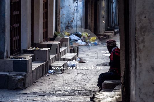 At dawn a Muslim man rests on a step in a narrow alley waiting for shops to begin trading. Stone Town, Mji Mkongwe, Zanzibar, Unguja Island, Zanzibar, Tanzania.