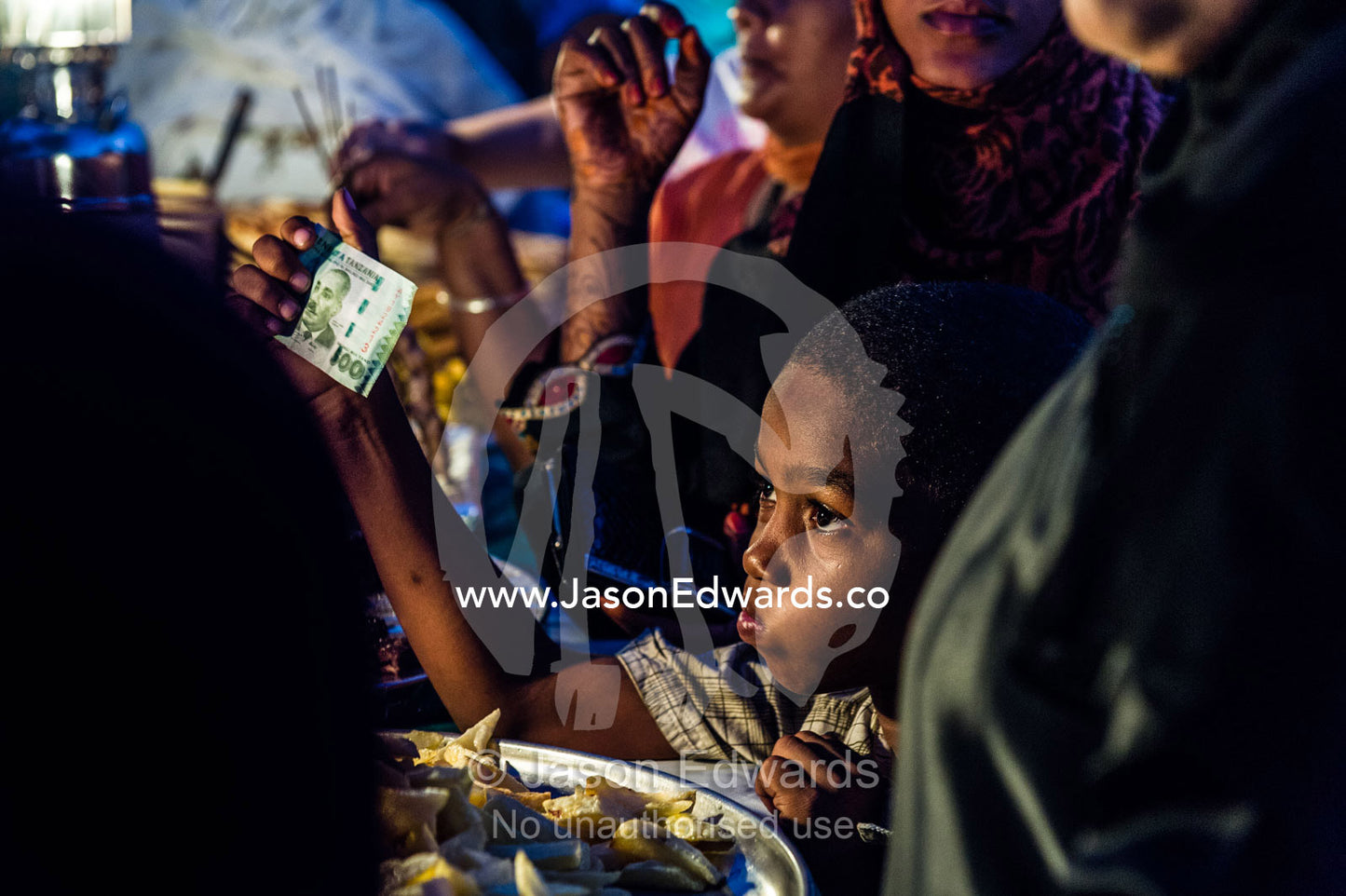A boy in a crowd waving money at a stall owner to buy food at a night market by the Indian Ocean. Forodhani Gardens, Stone Town, Mji Mkongwe, Zanzibar, Unguja Island, Zanzibar, Tanzania.