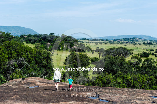 A mother and son hiking across the top of a granite kopje above a savannah plain. Moru Kopjes, Serengeti National Park, Serengeti, World Heritage Site, Tanzania.