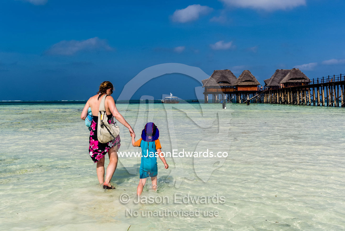 A mother and son wade across clear sandy shallows towards a dive boat. Bwejuu, Blue Lagoon Reef, Unguja Island, Zanzibar, Tanzania.