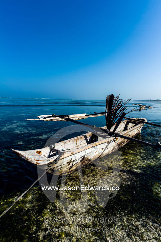 A wooden trimaran fishing dhow resting in the shallows of a tropical island at low tide. Bwejuu, Echo Beach Hotel, Echo Beach, Unguja Island, Zanzibar, Tanzania.