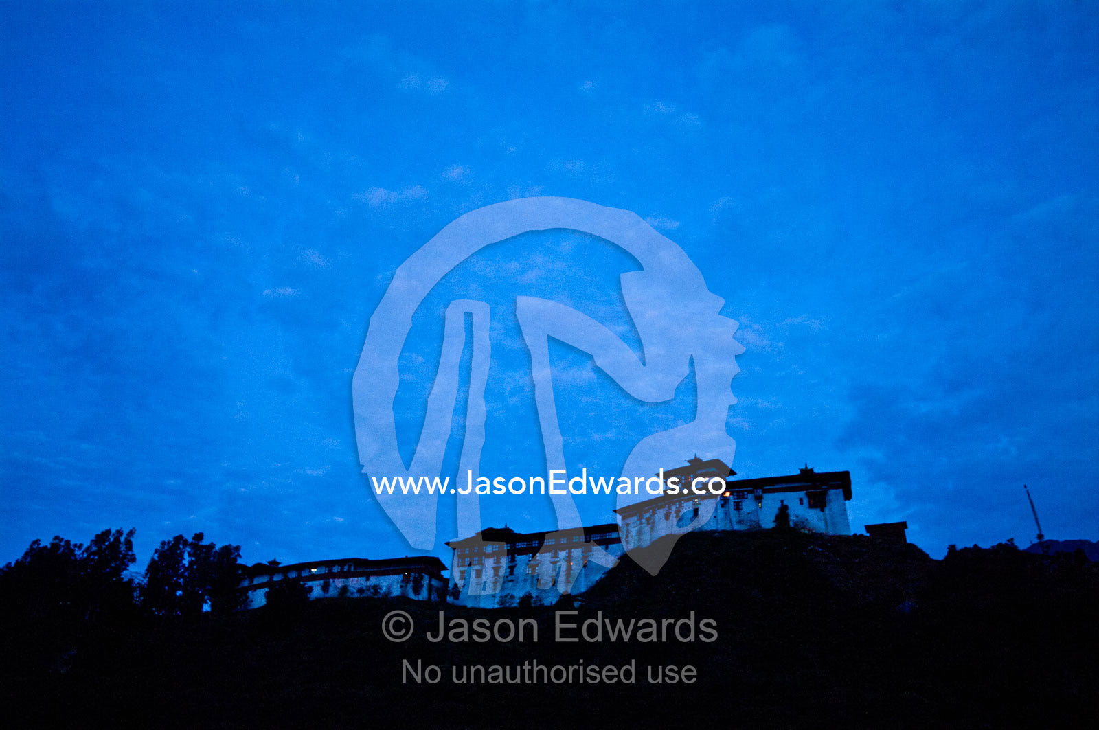 Lamp light peeks from windows atop the high walls of an ancient Buddhist monastery on a mountaintop at night. Punakha River, Punakha Valley, Punakha-Wangdiphodrang District, Bhutan.