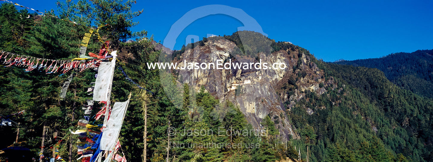 Prayer flags near an ancient Buddhist monastery perched on a sheer cliff face above a forested valley in the Himalaya. Paro Taktsang, The Tigers Nest, Paro Valley, Paro District, Bhutan.