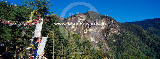 Prayer flags near an ancient Buddhist monastery perched on a sheer cliff face above a forested valley in the Himalaya. Paro Taktsang, The Tigers Nest, Paro Valley, Paro District, Bhutan.