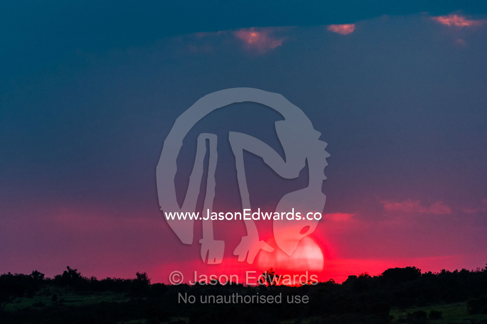 A blazing red sun saturated with dust kisses the horizon above the savannah plain at sunset. Maasai Mara National Reserve, Narok County, Kenya.