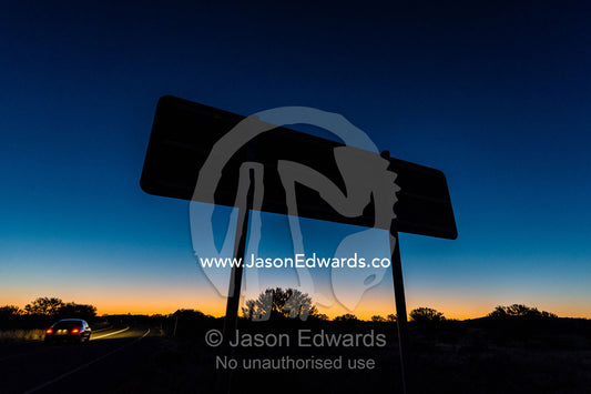 A car passes a directional sign silhouetted on a remote outback desert road at sunset. Uluru Kata Tjuta National Park, Northern Territory, Australia.