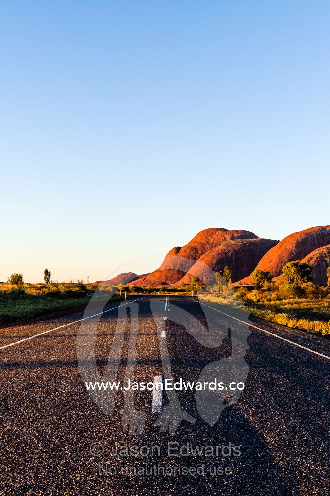 A bitumen road crosses the outback desert plain towards the domes of Kata Tjuta at sunset. Uluru Kata Tjuta National Park, Northern Territory, Australia.