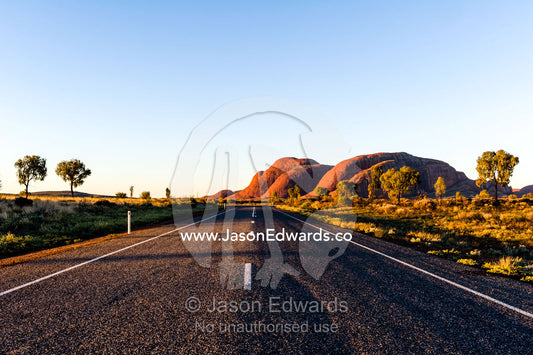 A bitumen road crosses the outback desert plain towards the domes of Kata Tjuta at sunset. Uluru Kata Tjuta National Park, Northern Territory, Australia.