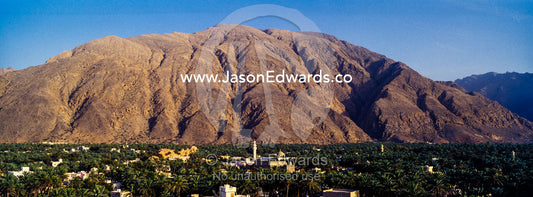 A date palm tree grove and village fills a desert valley floor beneath jagged mountain peaks. Wilayt Nakhal, Mount Nakhal, Al Batinah South Governorate, Sultanate of Oman.