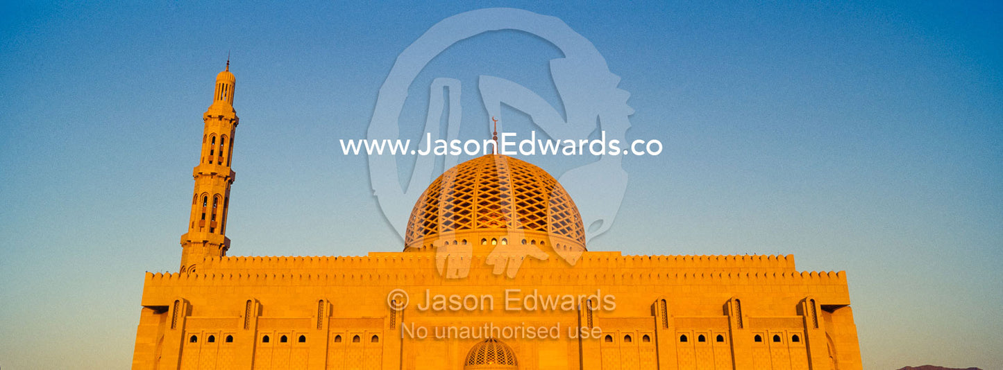 An ornate stone minaret towers over the dome of the Prayer Hall of the Grand Mosque at sunset. Sultan Qaboos Grand Mosque, Muscat, Sultanate of Oman.