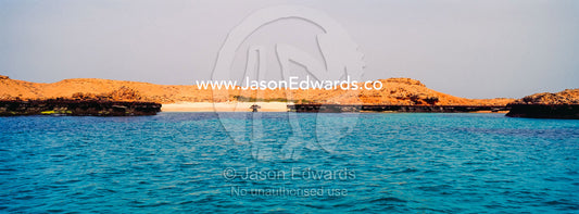 Clear, turquoise seas surround a barren red sandy desert island. Daymaniyat Islands Nature Reserve, Wilayat AlSeeb, Barka, Sultanate of Oman.