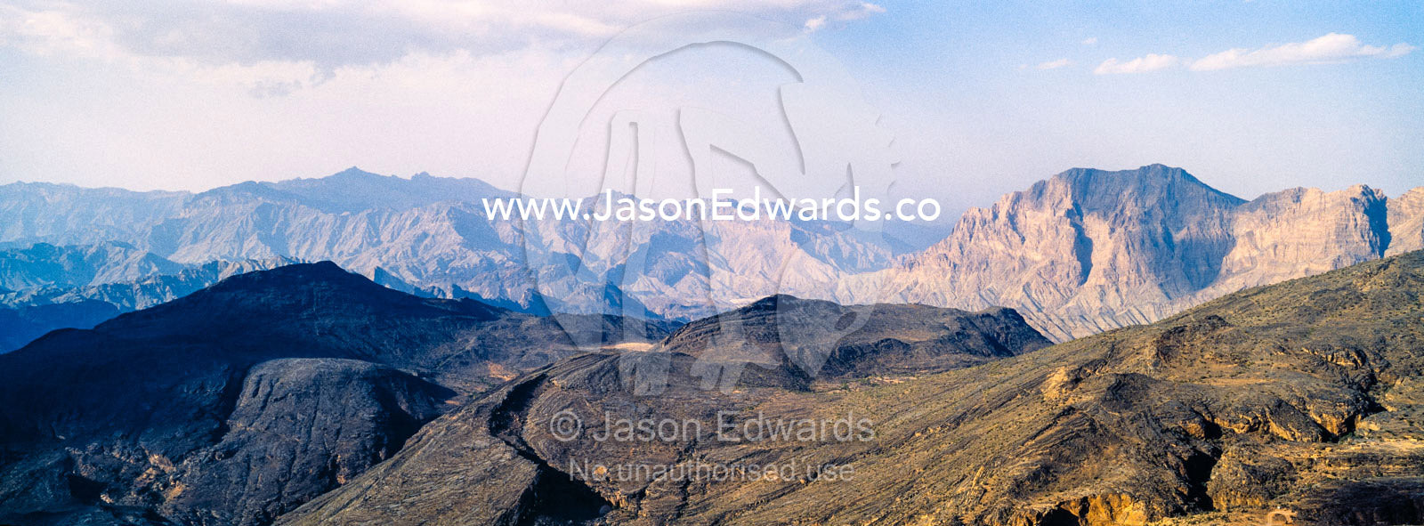 The steep slopes of arid jagged mountains overlooking an eroded and inhospitable rocky desert valley. Jebel Shams, Al Hajar Mountains, Sultanate of Oman.