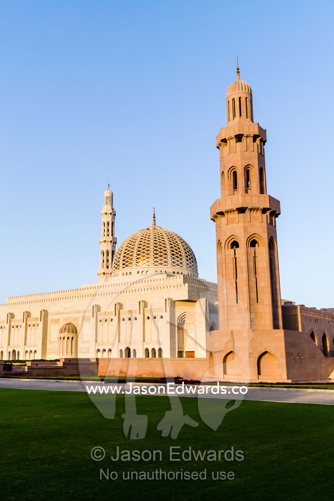An ornate stone minarette towers over the dome of the Prayer Hall of the Grand Mosque at sunset. Sultan Qaboos Grand Mosque, Muscat, Sultanate of Oman.