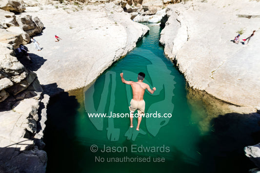 A tourist leaps into the turquoise waters of a wadi in a desert valley to cool off. Wadi Bani Khalid, Sharqiyah Region, Sultanate of Oman.