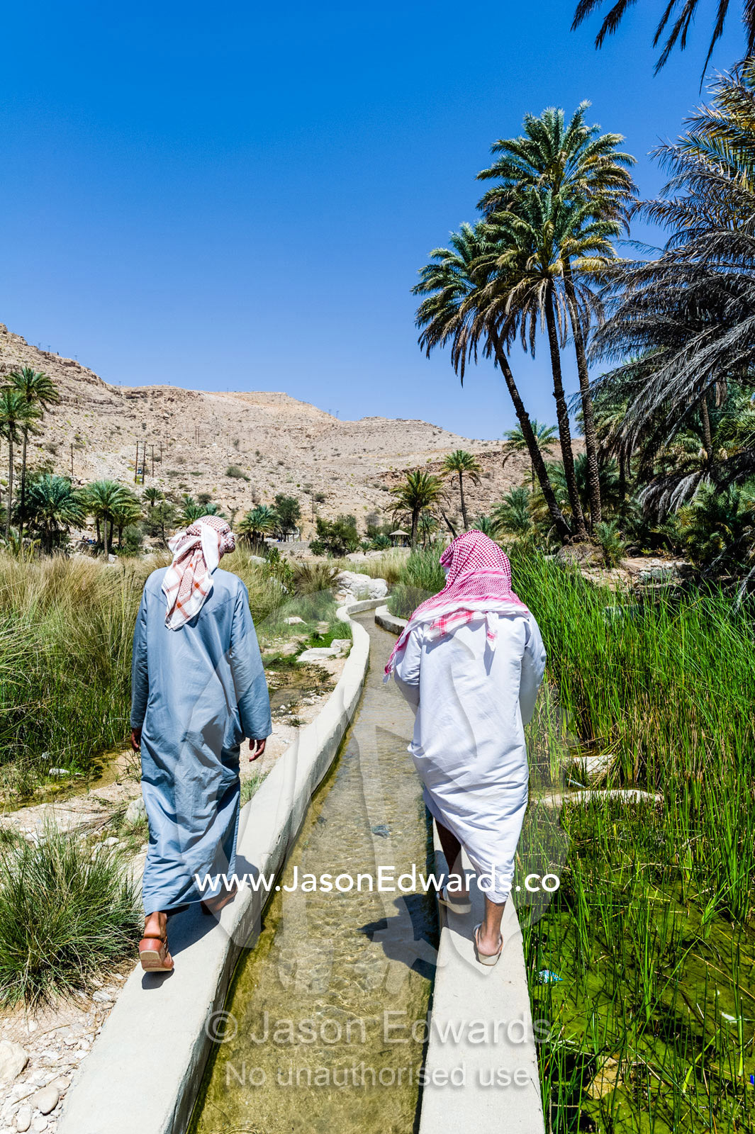 Arabic men wearing dishdasha and masar walk along a falaj transporting water from a desert wadi. Wadi Bani Khalid, Sharqiyah Region, Sultanate of Oman.