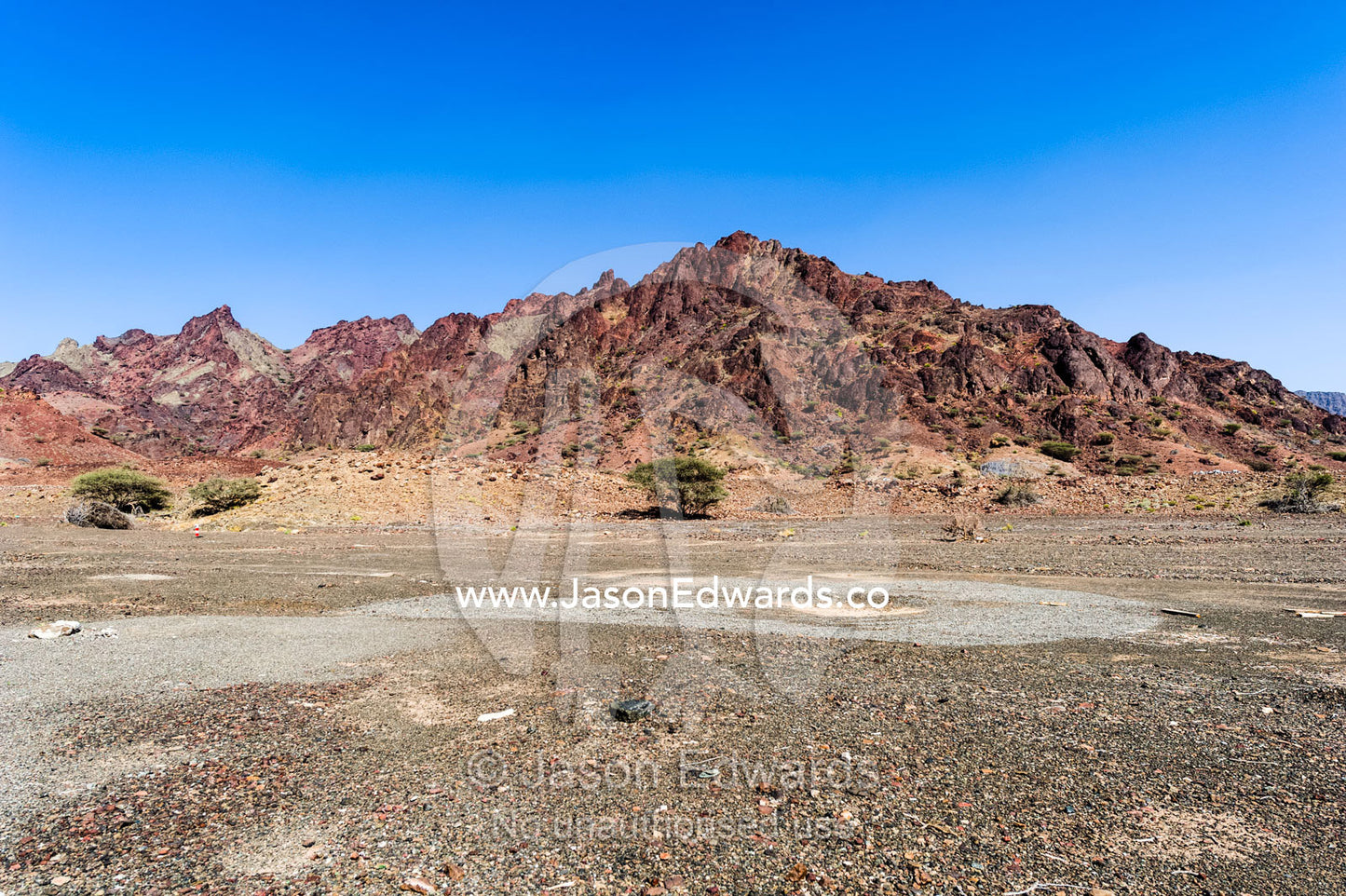 A red volcanic mountain rises from a gravel plain in the desert. Adh Dhahir, Sultanate of Oman.