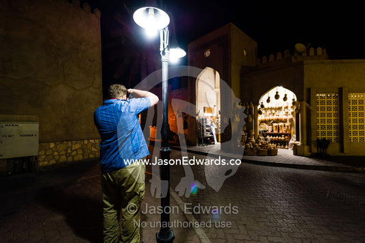 A photographer captures a store in a night market from beneath a street lamp. Mutrah Souq, Muscat, Sultanate of Oman.