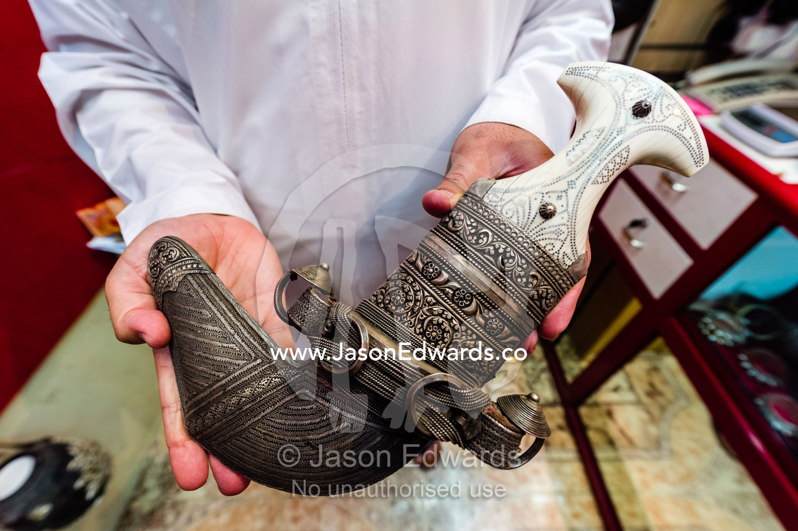 A salesman displays an expensive silver khanjar knif with an ivory handle for sale in a souq. Mutrah Souq, Muscat, Sultanate of Oman.
