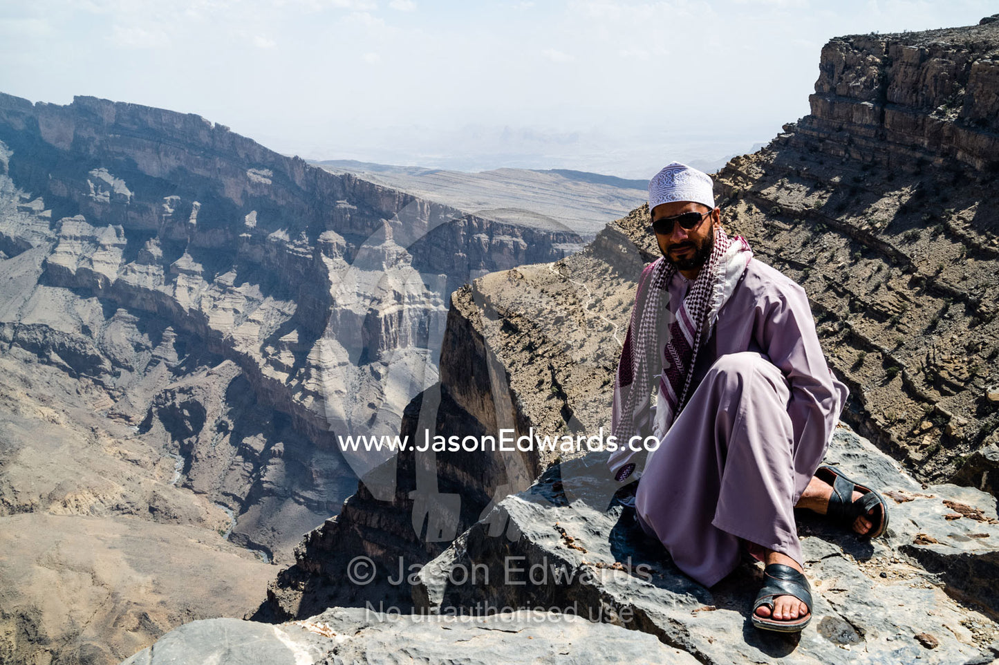 A guide sits beside sheer cliff walls and terraces carved by erosion in a massive desert canyon. Wadi Ghul, Jebel Shams, Al Hajar Mountains, Sultanate of Oman.