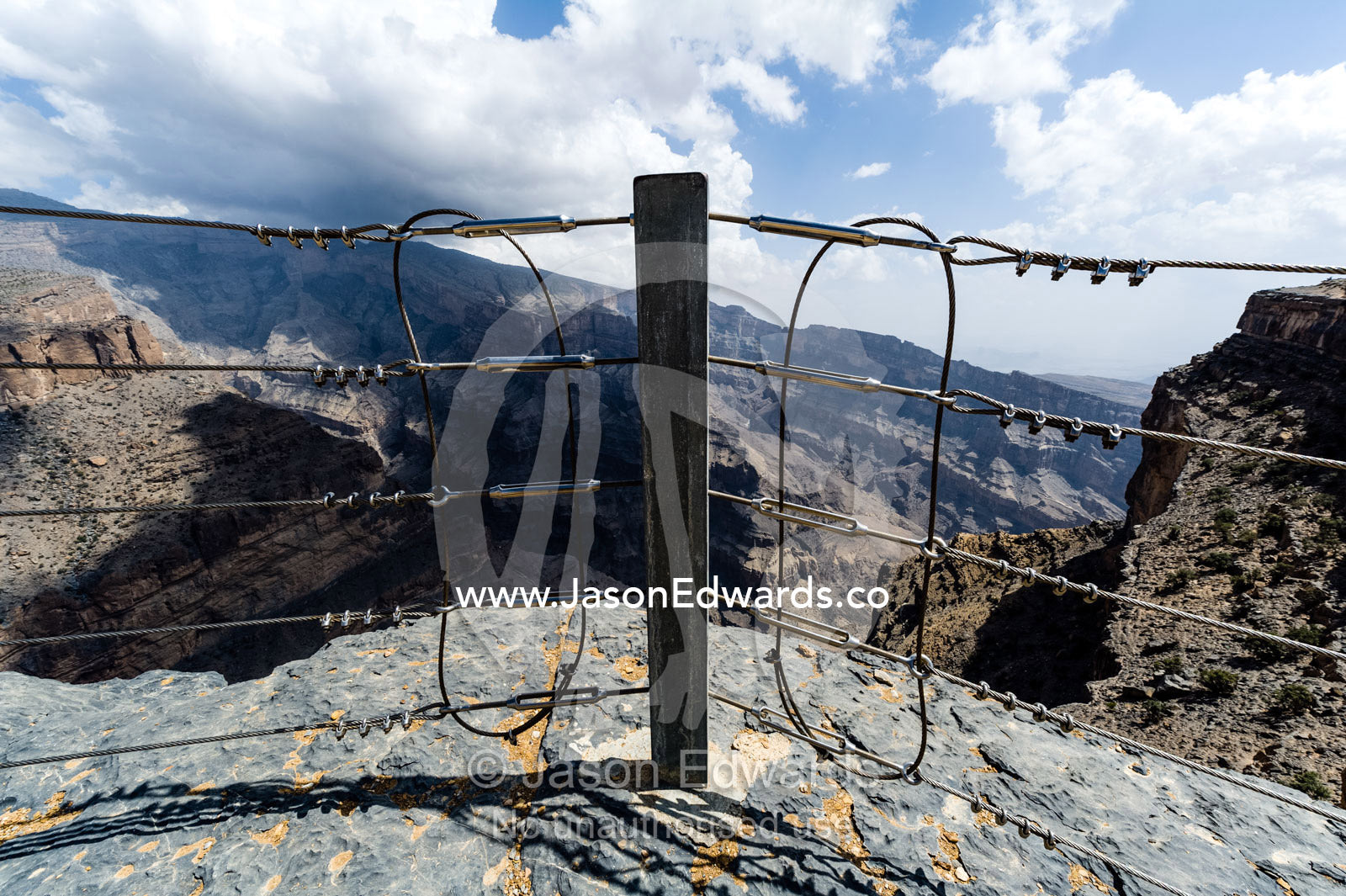 A safety fence near sheer cliff walls and terraces carved by erosion in a massive desert canyon. Wadi Ghul, Jebel Shams, Al Hajar Mountains, Sultanate of Oman.