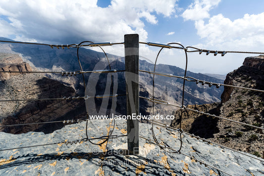 A safety fence near sheer cliff walls and terraces carved by erosion in a massive desert canyon. Wadi Ghul, Jebel Shams, Al Hajar Mountains, Sultanate of Oman.