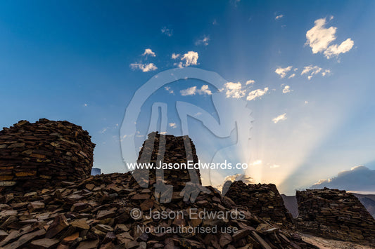 Dawn sun rays rise over ancient handmade stone burial mounds and tombs atop a desert ridge. Archaeological Sites of Bat, Al-Ayn, Al Dhahira Region, Sultanate of Oman.