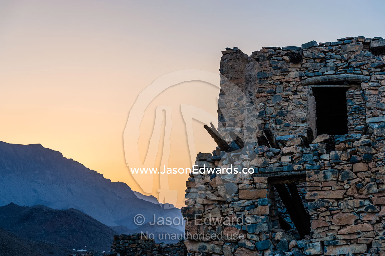 An ancient handmade stone building wall in an abandoned desert village overlooks the mountains at sunset. Wadi Nakhar, Al Hamra, Ad Dakhiliyah Region, Sultanate of Oman.