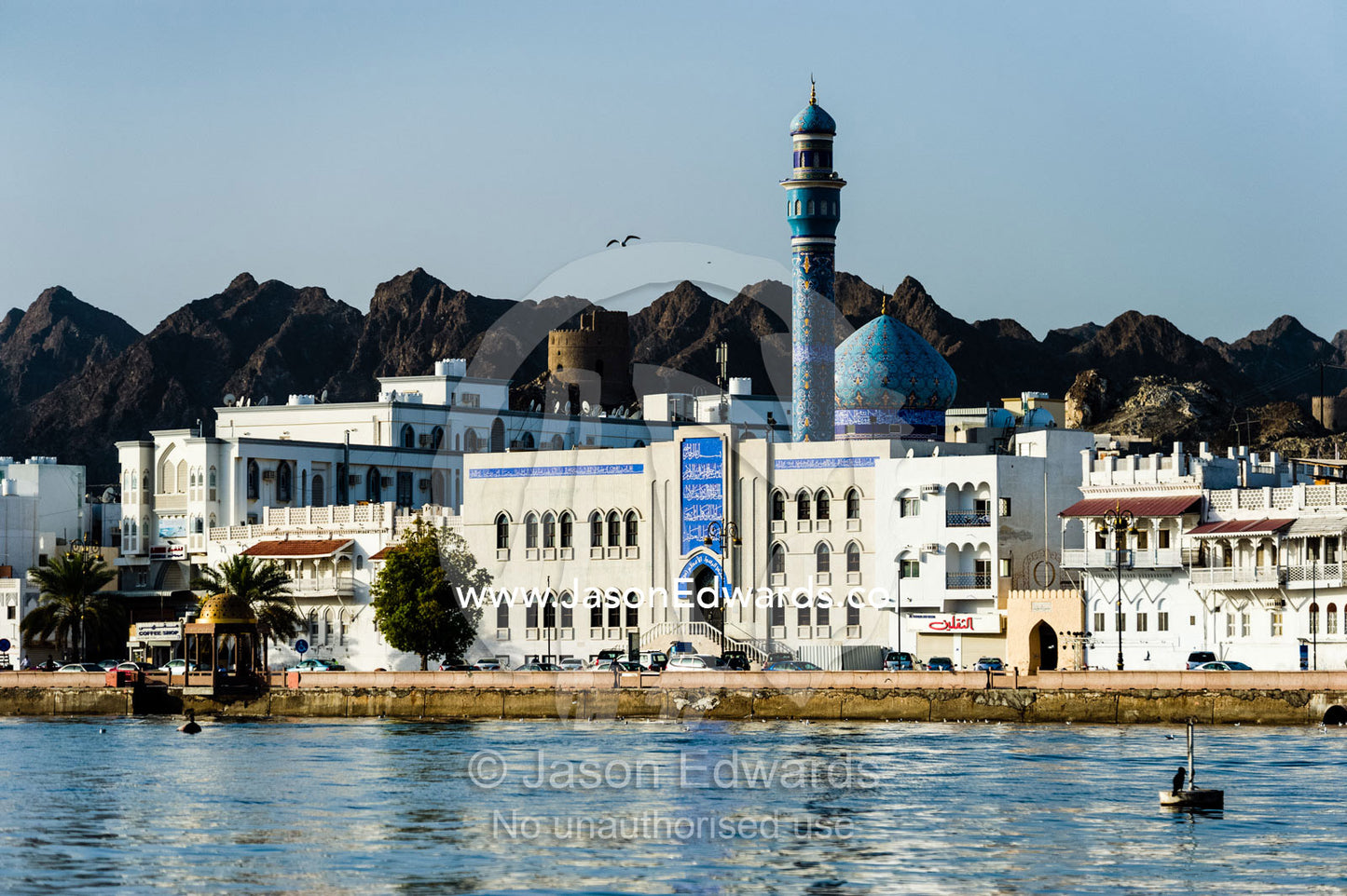 Desert mountains tower over whitewashed walls of a mosque and minaret behind a shopping and hotel district on the shores of the Gulf of Oman. Muscat, Gulf of Oman, Sultanate of Oman.
