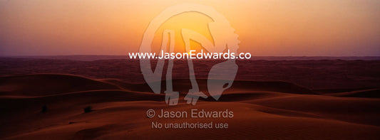 The sun dips below the distant horizon over a vast field of red sand dunes. Wahiba Sands, Ramlat al-Wahiba, Sharqiya Sands, Sultanate of Oman.
