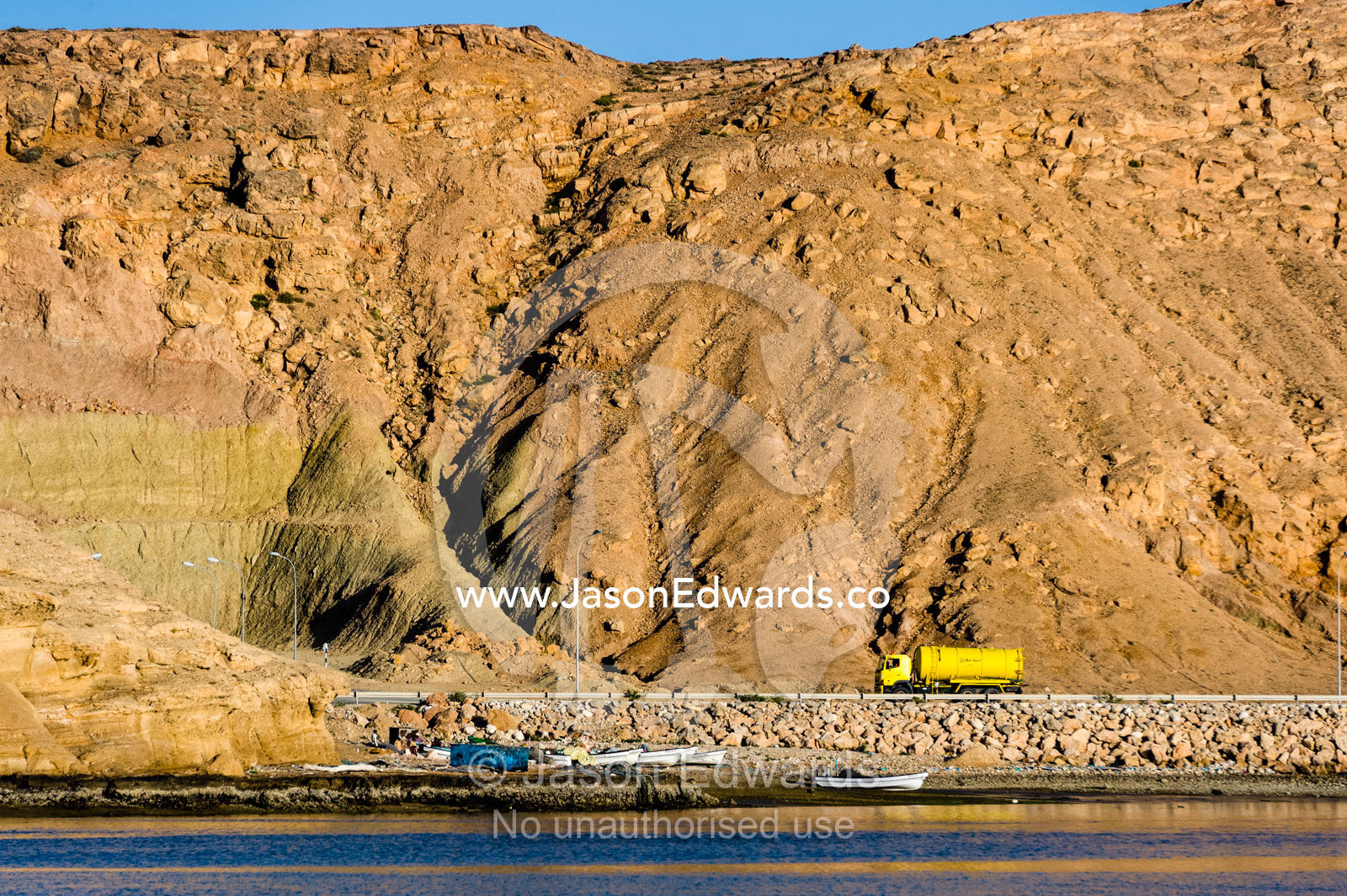 A tanker truck driving beneath a desert cliff along the coast of the Gulf of Oman. Sur, Ash Sharqiyah Region, Gulf of Oman, Sultanate of Oman.