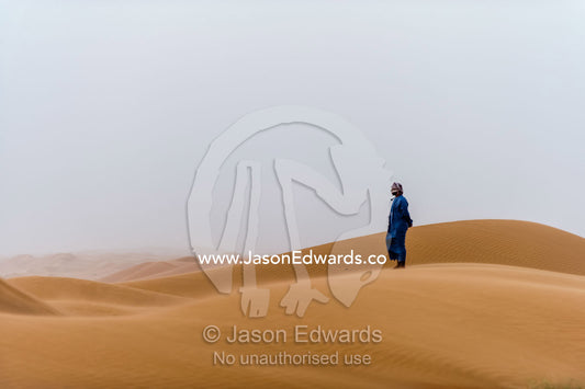 A Muslim man standing on a sand dune at sunset wearing a traditional dishdasha and masar. Wahiba Sands, Ramlat al-Wahiba, Sharqiya Sands, Sultanate of Oman.