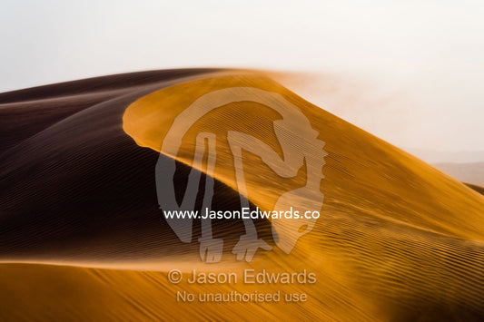 Sunset caressing the crests of red sand dune waves and howling wind in the desert. Wahiba Sands, Ramlat al-Wahiba, Sharqiya Sands, Sultanate of Oman.