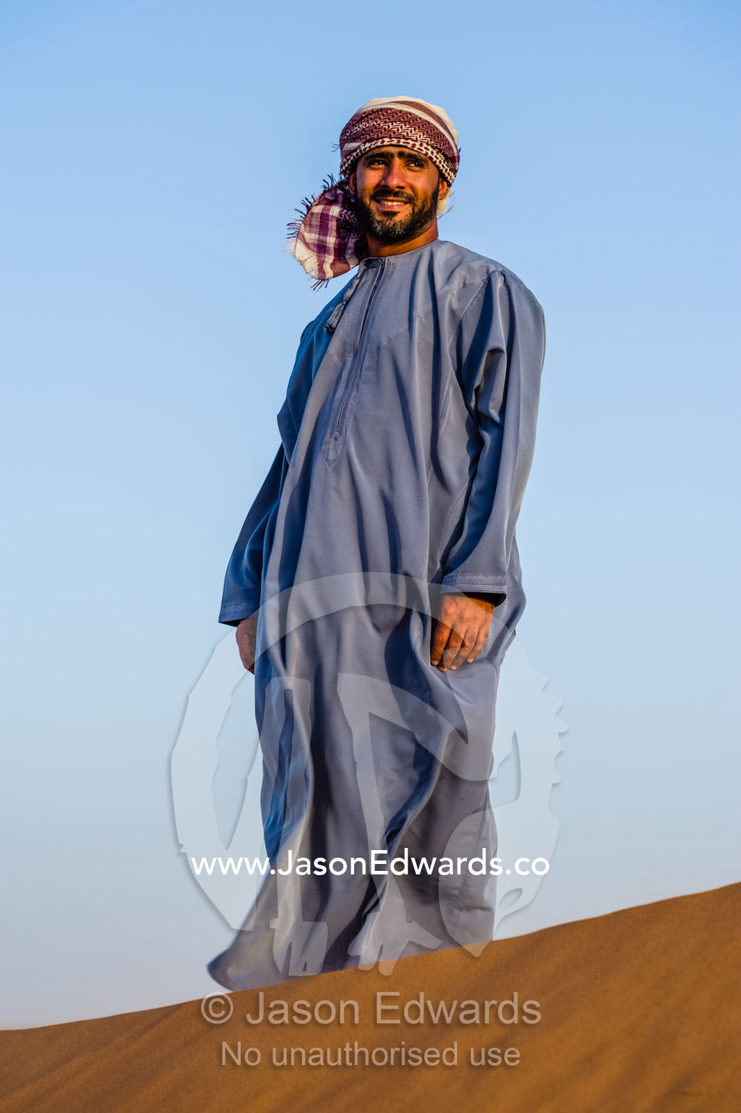 A Muslim man standing on a sand dune at sunset wearing a traditional dishdasha and masar. Wahiba Sands, Ramlat al-Wahiba, Sharqiya Sands, Sultanate of Oman.