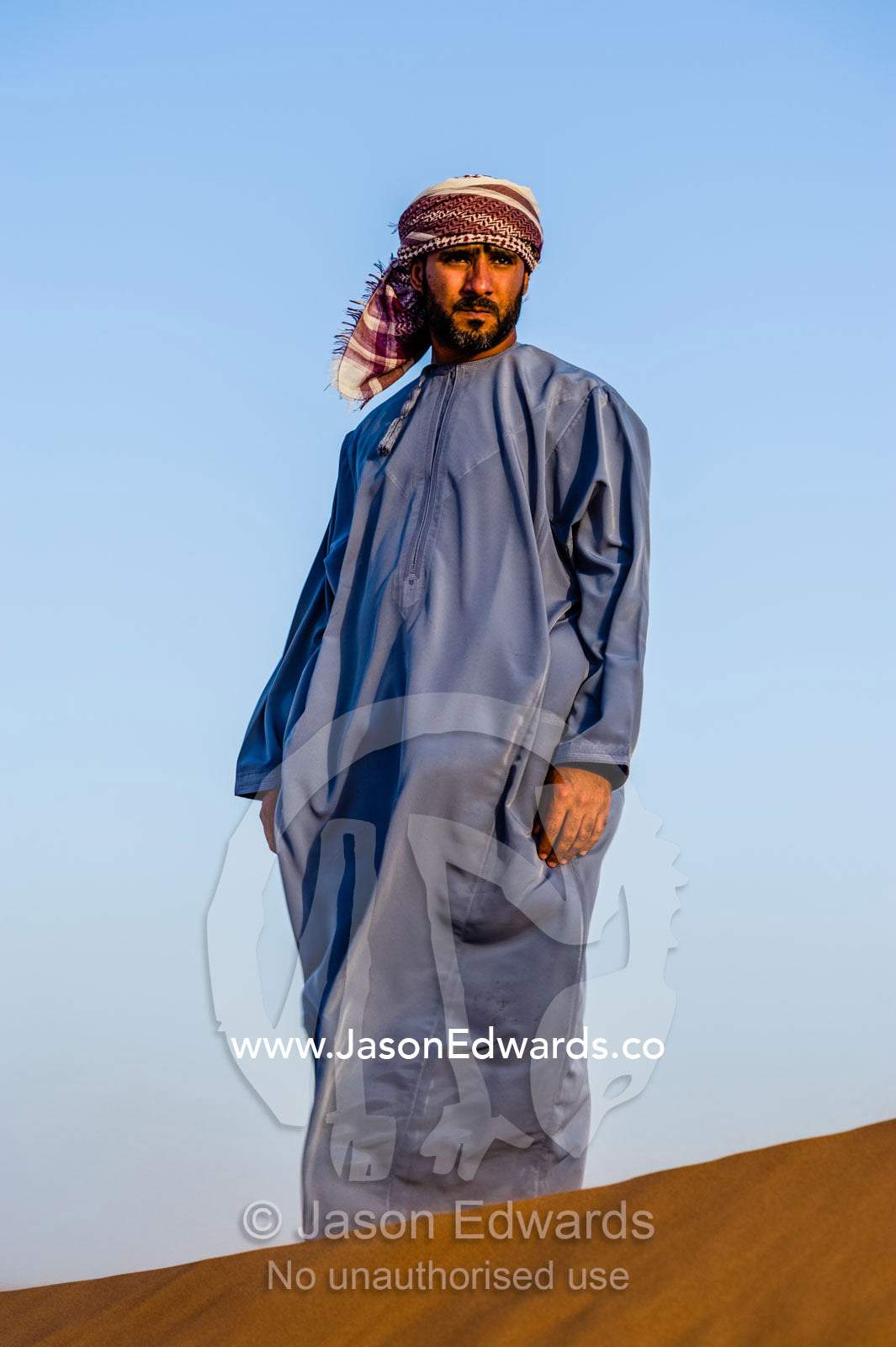 A Muslim man standing on a sand dune at sunset wearing a traditional dishdasha and masar. Wahiba Sands, Ramlat al-Wahiba, Sharqiya Sands, Sultanate of Oman.