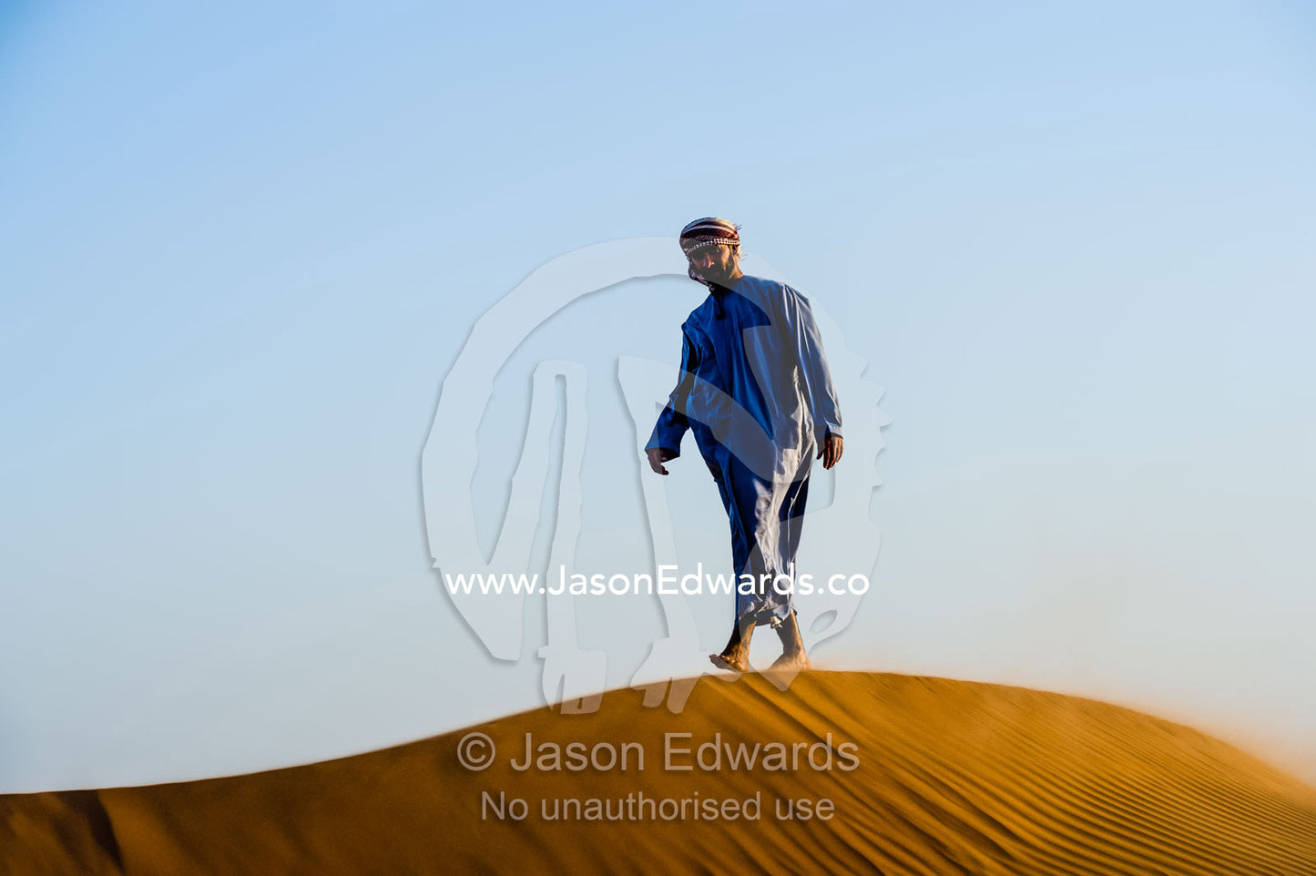 A Muslim man standing on a sand dune at sunset wearing a traditional dishdasha and masar. Wahiba Sands, Ramlat al-Wahiba, Sharqiya Sands, Sultanate of Oman.
