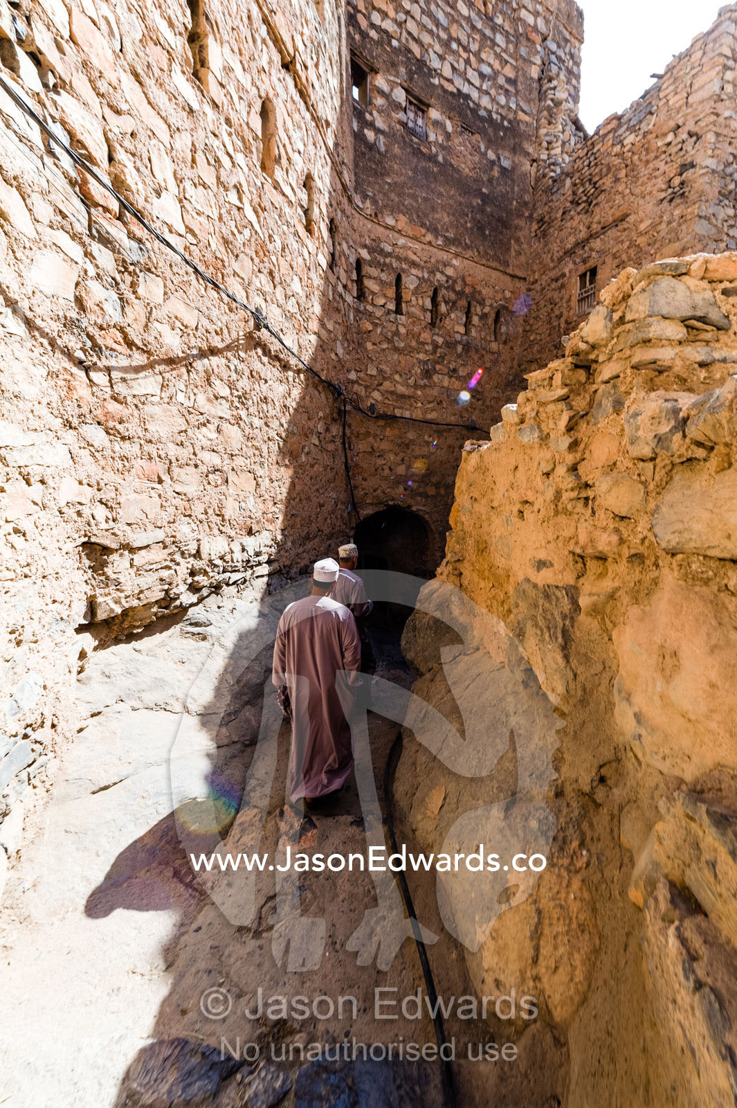 Muslim men walking along a narrow mud brick alleyway in an ancient village in the desert. Misfat Al Abryeen, Al Hajar Mountains, Al Hamra, Ad Dakhiliyah Region, Sultanate of Oman.