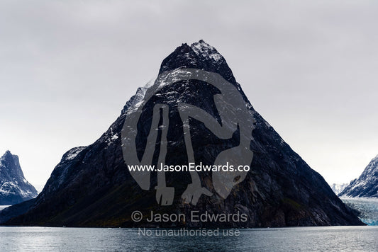 A cone-shaped mountain rises from the dark waters of an Arctic fjord beneath a storm laden sky. Fjord of Eternity, Maniitsoq, Sukkertoppen, Qeqqata Municipality, Greenland.