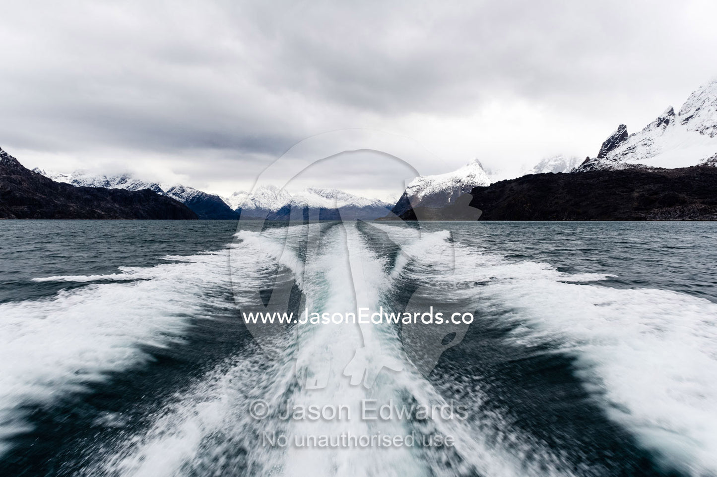 A boats wake peels between snow covered peaks in a fjord. Fjord of Eternity, Maniitsoq, Sukkertoppen, Qeqqata Municipality, Greenland.