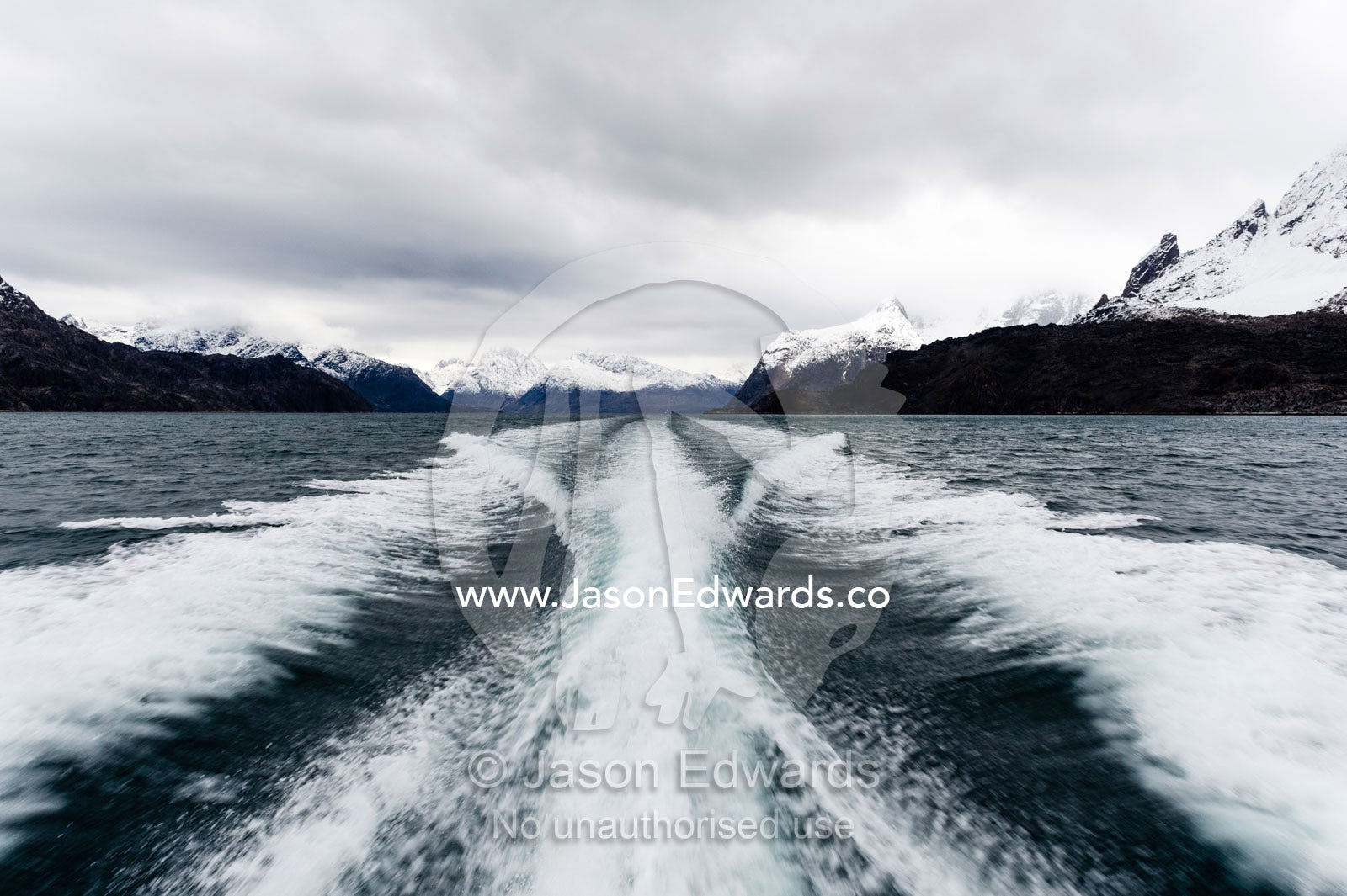 A boats wake peels between snow covered peaks in a fjord. Fjord of Eternity, Maniitsoq, Sukkertoppen, Qeqqata Municipality, Greenland.