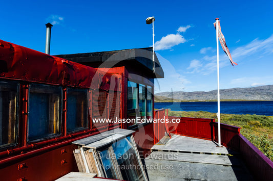 A red cottage with spare window frames overlooks an alpine lake on a sunny morning. Tasersuatsiaq Lake, Kangerlussuaq, Qeqqata Municipality, Greenland.
