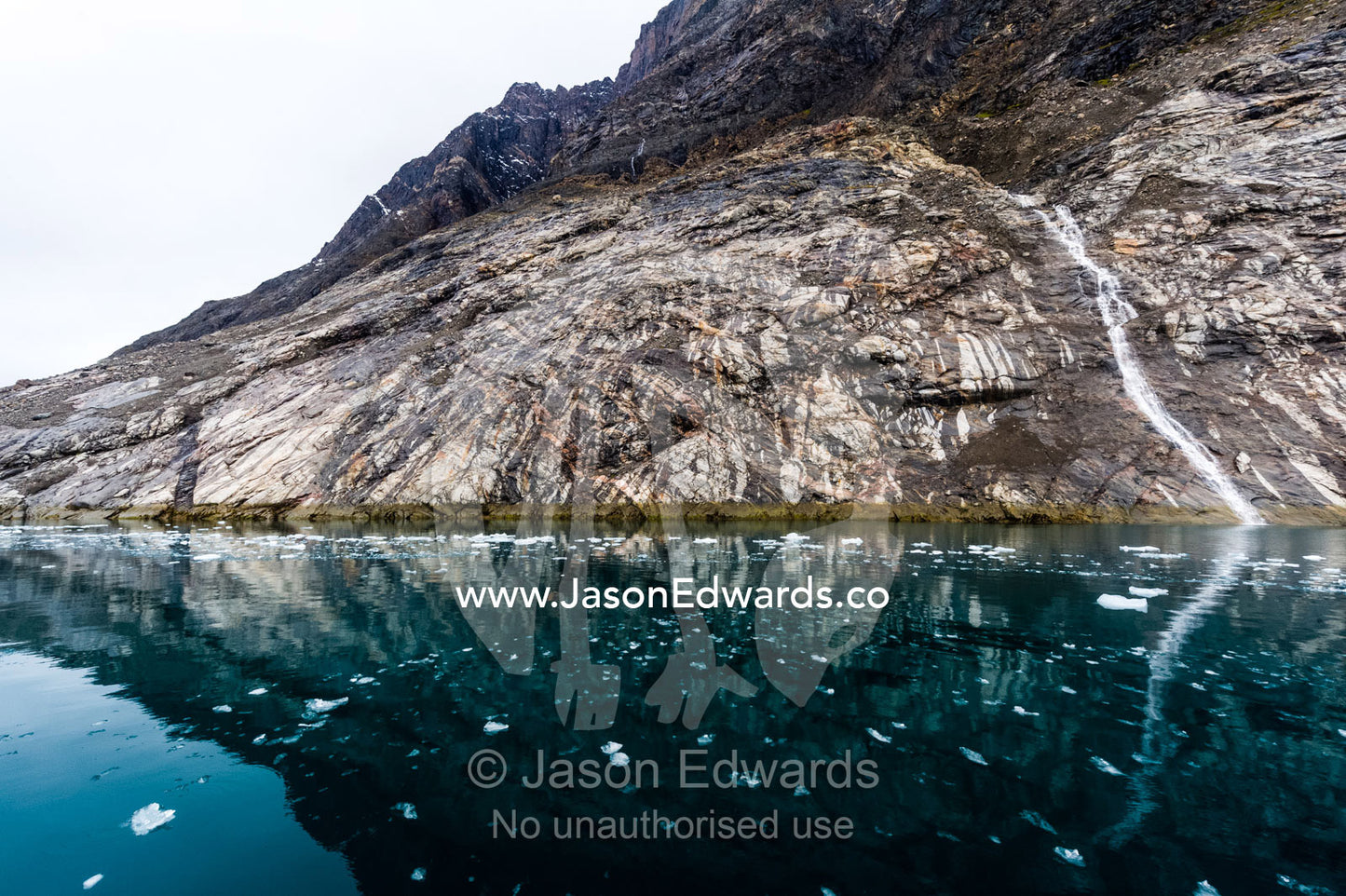 A small waterfall running down a cliff into the darks waters of a fiord. Fjord of Eternity, Maniitsoq, Sukkertoppen, Qeqqata Municipality, Greenland.