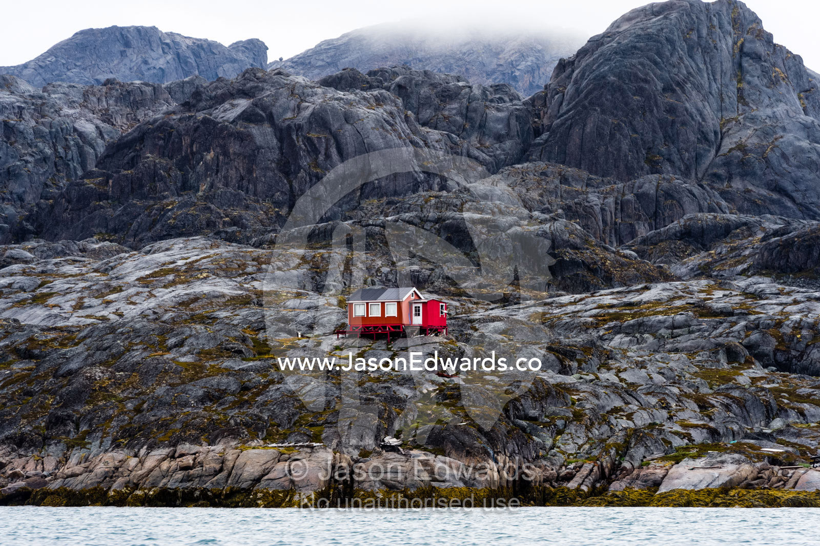 A bright red cottage on the rugged and brutal rock shoreline of a fjord. Davis Strait, Maniitsoq, Sukkertoppen, Qeqqata Municipality, Greenland.