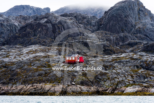 A bright red cottage on the rugged and brutal rock shoreline of a fjord. Davis Strait, Maniitsoq, Sukkertoppen, Qeqqata Municipality, Greenland.