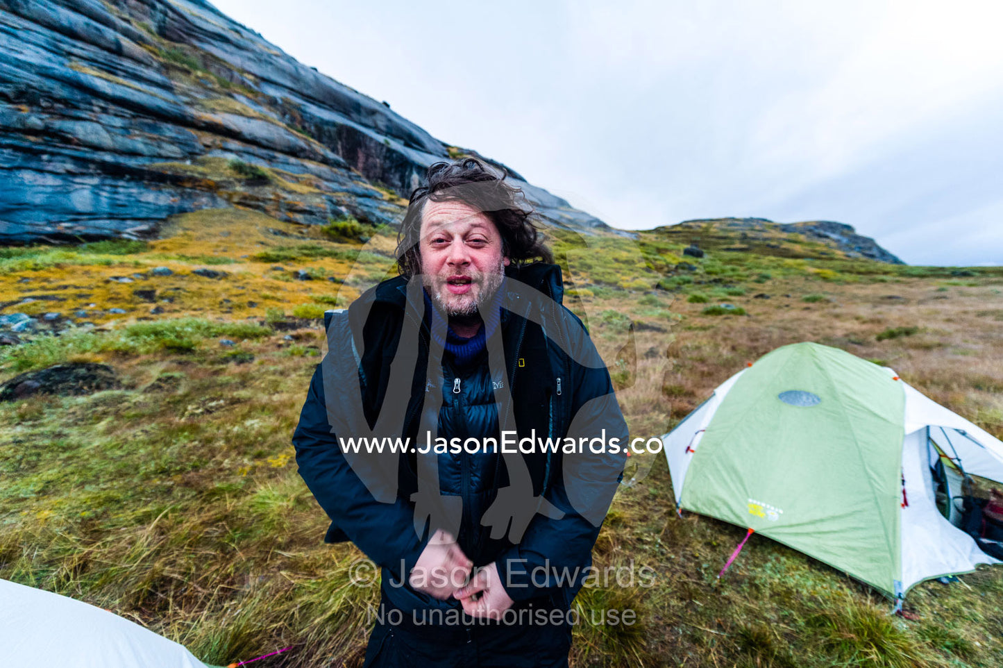 A hiker zips up his jacket to stay warm from the bitter wind after leaving his tent. Ammalortup Nunaa Highland, Kangerlussuaq, Qeqqata Municipality, Greenland.