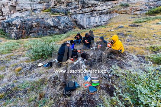A group of hikers prepare a meal beneath a hill away from the chilly wind. Ammalortup Nunaa Highland, Kangerlussuaq, Qeqqata Municipality, Greenland.