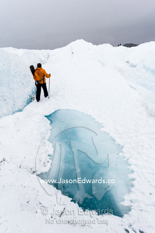 A hiker passing between a dangerous crevasse and a melt pond on the Greenland Ice Shelf. Greenland Ice Sheet, Qeqqata Municipality, Kangerlussuaq, Greenland.