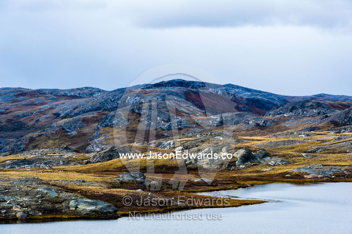 Outcrops of exposed barren rocks emerge through the tundra surrounding an alpine lake. Isunngua Highland, Kangerlussuaq, Qeqqata Municipality, Greenland.